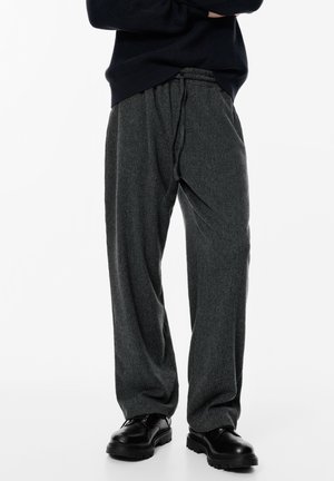 Személy laza, sötétszürke kötött nadrágot visel, amit zsinórral lehet összehúzni, fekete, vastag talpú cipővel és fekete pulóverrel, karba tett kézzel.