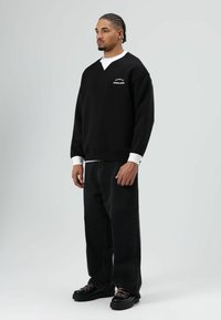 Zwarte oversized sweatshirt met een V-hals en witte stofaccenten bij de manchetten, gecombineerd met zwarte wijd uitlopende jeans en chunky zwarte schoenen.