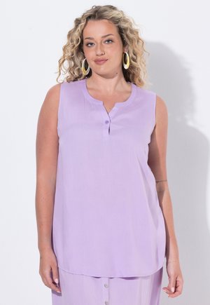Donna con capelli biondi ricci che indossa grandi orecchini a cerchio e una canotta viola chiaro con bottoni su sfondo semplice.
