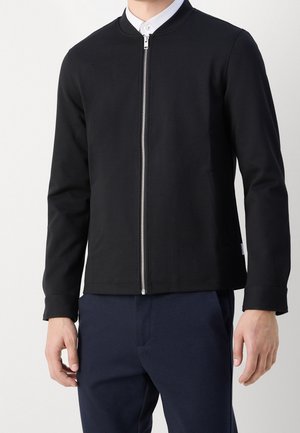 Homme portant une veste noire zippée sur une chemise blanche à col et un pantalon bleu foncé, debout devant un fond uni.