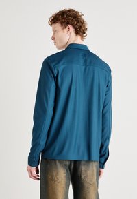 Teal long-sleeve shirt with vertical ribbing, a spread collar, and buttoned cuffs. The back features a yoke design and a smooth texture.