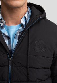 Giacca a vento nera con cappuccio, dotata di chiusura a zip e zip di colore blu. Logo ricamato sul petto e camicia a quadri sovrapposta.