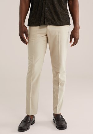 Homme portant un pantalon beige sur mesure, des mocassins noirs et une chemise sombre boutonnée, debout devant un fond neutre.