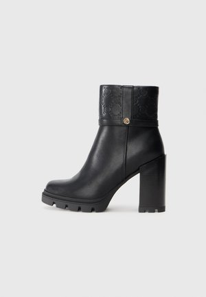 Botte noire en cuir jusqu'à la cheville avec un talon épais, dotée d'un bandeau supérieur texturé, de motifs en relief et d'un accent doré rond.