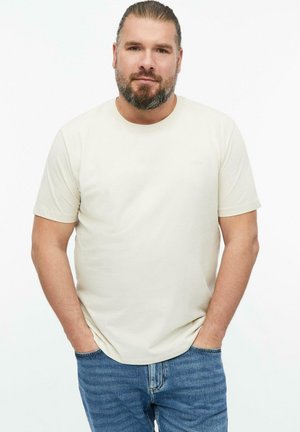 Man with beard and tied-back hair stands with hands in pockets wearing a plain light beige t-shirt and blue jeans against white background.