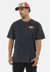 T-shirt à manches courtes bleu marine foncé avec un logo sur la poitrine, fabriqué en tissu doux. Le porteur porte également une casquette multicolore et un pantalon noir.