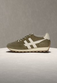 Baskets en daim vert olive avec une bande latérale blanche, un talon blanc, des lacets à motifs et une semelle en caoutchouc gomme sur une surface claire et un fond neutre.