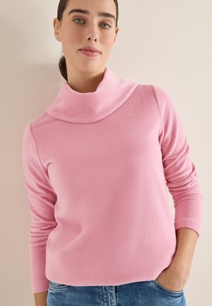 Kvinna som bär en rosa långärmad tröja med rullad polokrage och blå jeans, stående mot en ljusvägg.