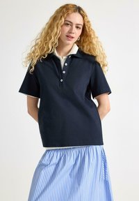 Marineblå polo shirt med hvid krave, der har en knappelukning og fem knapper. Kombineret med en lyseblå stribet nederdel.
