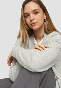 Hellgrauer Cardigan mit gerippten Bündchen, lässiger Schnitt, mit einem Knopfakzent. Getragen über einem weißen Hemd, kombiniert mit dunkelgrauen Hosen.