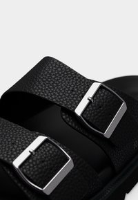 Μαύρα δερμάτινα σανδάλια με δύο φαρδιές λουρίδες που διαθέτουν ασημί ορθογώνιες αγκράφες, υφή στην επιφάνεια και λεία μαύρη σόλα.