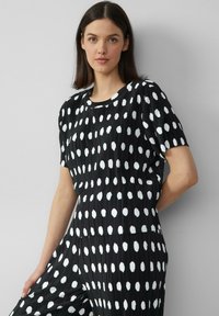 Zwarte geplooide jumpsuit met witte stippen, met een ronde halslijn en korte mouwen. De textuur is soepel met een lichte glans.