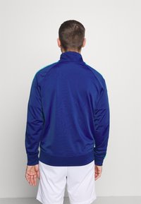 Giacca sportiva blu con colletto alto, maniche allungate e dettagli azzurri sulle spalle. Tessuto liscio e design aderente.