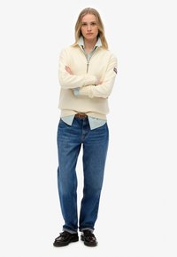 Crème trui met een hoge hals en rits, gelaagd over een blauwgestreept shirt. Gecombineerd met losse blauwe jeans en donkere schoenen. Eenvoudige stijl.