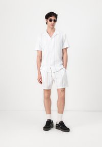 Kortærmet skjorte med krave og matchende shorts i hvid med lyseblå striber. Båret med sorte sko, hvide sokker og solbriller.