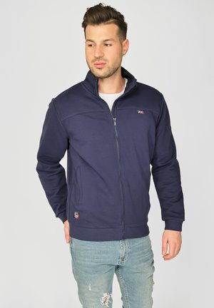 Marineblå zip-up jakke i blødt stof med høj krave, har sidelommer, logo på brystet og ribbede manchetter og kant.