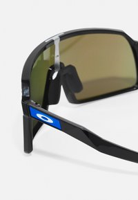 Gafas de sol deportivas con un marco negro brillante, lentes verdes y un acento azul en la patilla. Incluyen un agarre de goma para mayor estabilidad.