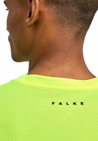 Jasnożółta sportowa koszulka z siateczki z okrągłym dekoltem i czarnym logo "FALKE" na plecach. Tkanina ma przewiewną strukturę.