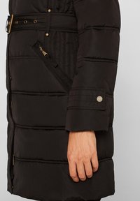 Vue latérale d'une personne portant un manteau matelassé noir avec une taille ceinturée, une poche zippée en biais et un détail de manche à boutons-pression.