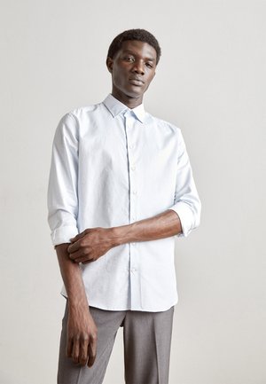 Camisa de botones azul claro con un patrón sutil, mangas largas arremangadas en los puños, combinada con pantalones grises. Diseño casual y a medida.
