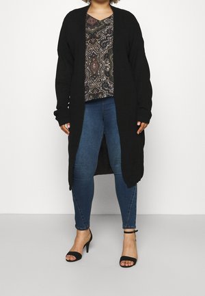 Femme portant un long cardigan noir, un top à motifs, un jean skinny bleu et des talons ouverts noirs avec des brides à la cheville, debout sur un fond blanc.
