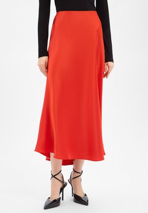 Femme portant une jupe midi asymétrique rouge vif, un haut noir à manches longues et des chaussures à talons hauts noires à brides, debout sur un fond blanc.