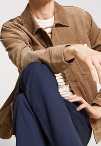 Råge jacka i ljusbrun, med krage, framknappar och sidofickor. Bärs över en randig skjorta och marinblå byxor.