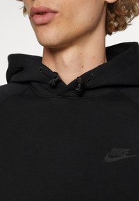 Svart Nike-hoodie med dragsko i huvan, slät textur och en diskret logotyp på bröstet. Närbild på halsringningen och ansiktet.