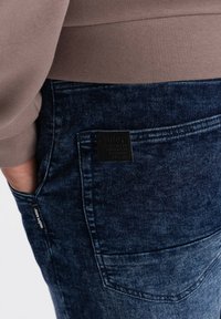Jeans en denim bleu foncé avec une finition texturée. Présente une étiquette en cuir noir sur la poche arrière et une étiquette de tissu visible sur le côté.