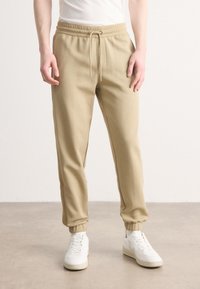 Beige joggebukser med elastisk midje og justerbar snor. Ankelmanchetter og en jevn tekstur, kombinert med hvite sneakers.
