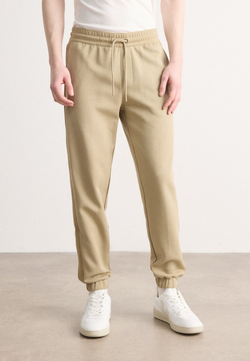 Beige joggebukser med elastisk midje og justerbar snor. Ankelmanchetter og en jevn tekstur, kombinert med hvite sneakers.