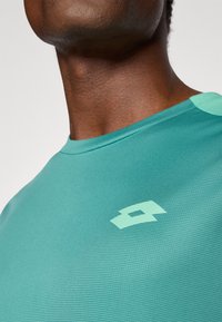 Tirkizna sportska majica s teksturiranom površinom i svijetlozelenim logotipom na prsima. Ima okrugli izrez i kratke rukave.