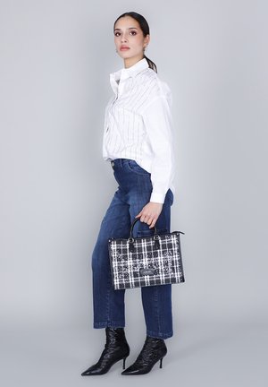 Borsa a tracolla a scacchi bianco e nero con manici doppi. Modella in camicia bianca con righe verticali e jeans blu, indossando stivali neri a punta.