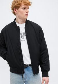 Νέος άντρας με σγουρά κόκκινα μαλλιά, φοράει μαύρο μπουφάν με φερμουάρ πάνω από λευκό μπλουζάκι με γραφικό σχέδιο και ανοιχτόχρωμο τζιν, κοιτάζει στο πλάι.