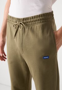 Olive spalvos sportinės kelnės su elastinga juosmens juosta, raištelių uždarymu ir šoninėmis kišenėmis. Kišenėje yra mažas mėlynas "HUGO" logotipas.