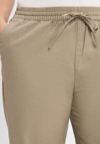 Beige joggerbroek van zacht materiaal, met een elastische tailleband en een trekkoord, zijzakken en een relaxed fit ontwerp.