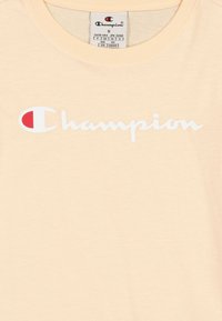 Camiseta de algodón de color beige claro con un logo en cursiva "Champion" en blanco y un círculo de acento rojo. Diseño clásico de cuello redondo, textura suave.