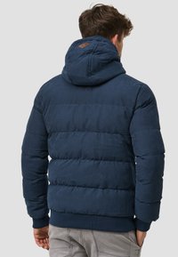 Veste rembourrée bleu marine avec une capuche, dotée d'une texture matelassée, de poignets côtelés et d'un badge en cuir à l'arrière.