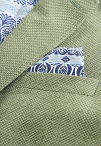 Blazer vert texturé avec un mouchoir de poche à motifs blanc, bleu et orange soigneusement rangé dans la poche poitrine.