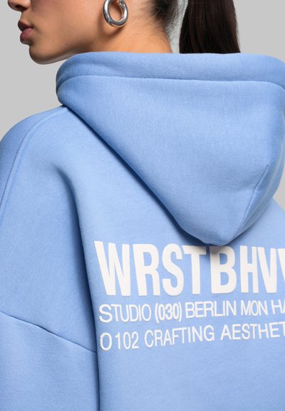 Jeune femme portant un sweat à capuche bleu avec le texte blanc WRSTBHV et une boucle d’oreille argentée, vue de côté et de dos sur un fond gris.