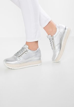 Stopy w metalicznych srebrnych sneakersach z bocznymi zamkami i sznurowadłami, połączone z białymi spodniami na jednolitym białym tle.