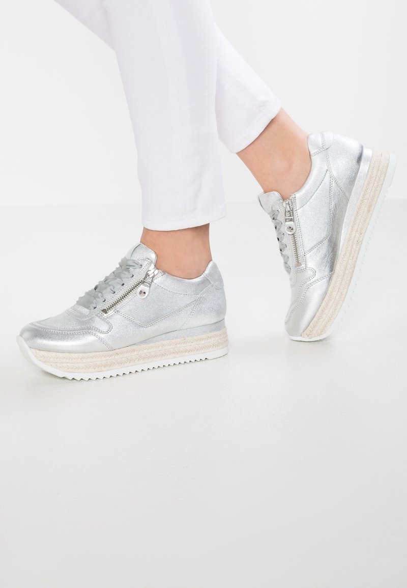Pieds portant des baskets argent métallisé avec des fermetures éclair latérales et des lacets, associés à un pantalon blanc sur un fond blanc uni.