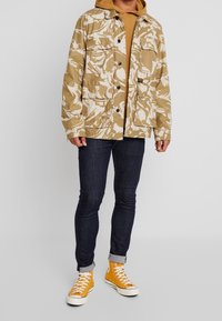 Chaqueta beige con patrón de camuflaje, cuatro bolsillos, cierre de botón, superpuesta sobre una sudadera marrón, combinada con jeans oscuros y zapatillas amarillas.