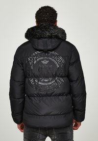Jacket puffer negru cu glugă căptușită cu blană, având un mare logo circulă embosat pe spate. Fabricată din material lucios, căptușit.