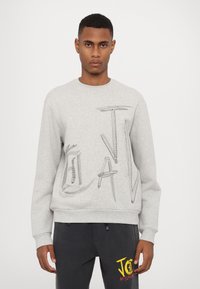Grå sweatshirt med broderad "JAV"-text i svart. Ribbad ärm och nederkant, borstad yta, rundad halsringning. Bärs med mörka byxor med en logotyp.