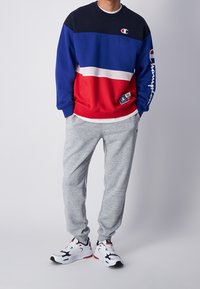 Rot, blau und marineblau farblich abgesetzter Pullover mit einem weißen Streifen, kombiniert mit grauen Jogginghosen und schwarz-weißen Sneakers.