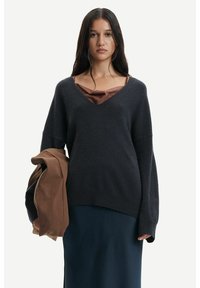 Maglione oversize grigio scuro con scollatura a V, maniche lunghe e tessuto strutturato. Indossato sopra un top marrone, abbinato a una gonna blu navy.