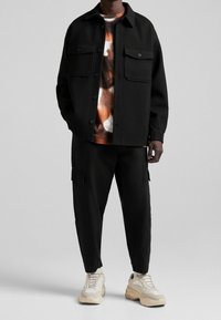 Černá kožená bunda s dvěma kapsami na hrudi, přes vzorovanou košili v hnědých a oranžových odstínech, doplněná černými cargo kalhotami a světlými teniskami.