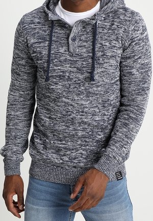 Uomo che indossa una felpa con cappuccio in maglia mélange blu e grigia con collo con bottoni e coulisse, abbinata a jeans azzurri.