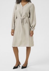 Femme portant une robe beige à manches longues, longueur genoux, avec une ceinture à nouer et des escarpins noirs à bout pointu, se tenant sur un fond blanc.
